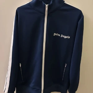 Mörkblå  Palm Angels S/M - Säljer en snygg mörkblå track jacket från Palm Angels med vita detaljer och dragkedja. Jackan har en hög krage och vita ränder längs ärmarna. Perfekt för en present till nån man tycker om.