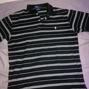 Svart och vit randig pikétröja från Ralph Lauren - Säljer en klassisk svart och vit randig pikétröja från Ralph Lauren. Tröjan har korta ärmar och en broderad logotyp på bröstet. Tillverkad i 100% Pima bomull för extra komfort. Perfekt för en stilren look!