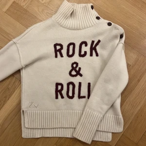 Vit stickad tröja från Zadig & Voltaire - Säljer en snygg vit stickad tröja från Zadig & Voltaire med texten 'Rock & Roll' i mörkröd på framsidan. Tröjan har en hög krage med knappar och ribbade detaljer vid ärmslut och nederkant. Perfekt för en cool och avslappnad stil. Tröjan saknar en knapp men inget man tänker på, utöver knappen som saknas finns det inga andra defekter. Hör av er vid intresse💞 pris kan diskuteras vid snabbt köp