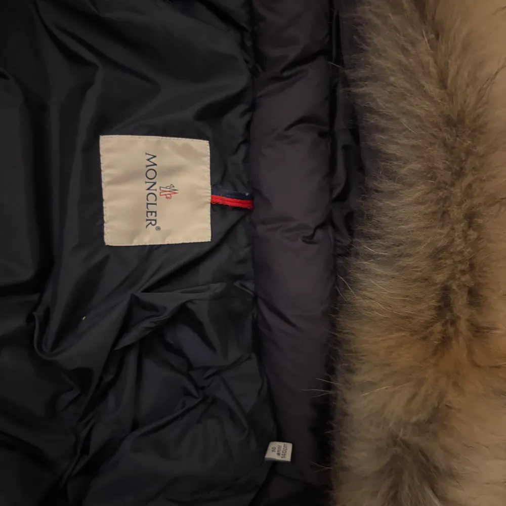 Snygg marinblå dunjacka från Moncler med en tjock lyxig pälskrage. Jackan har en dragkedja framtill och praktiska fickor med knappar. Perfekt för kalla vinterdagar. Två knappar saknas på luvan, men är lätt att åtgärda. . Takit.
