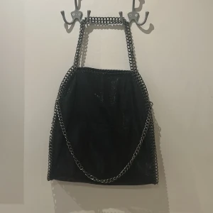 Svart axelväska med kedjedetaljer - Snygg svart axelväska med coola kedjedetaljer runt kanterna och som axelrem. Rymmer allt du behöver för dagen. Enkel att matcha med olika outfits. Säljer för har två stycken. Nyskick.