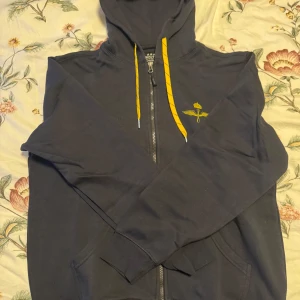 Mörkblå hoodie med Swedish Air Force-tryck - Snygg mörkblå hoodie med dragkedja och gult Swedish Air Force-tryck på ryggen. Den har en justerbar huva med gula snören och en liten logga på bröstet. Perfekt för en avslappnad stil.