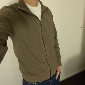 Cardigan från Massimo Dutti - Säljer en stilren beige tröja från Massimo Dutti med dragkedja och hög krage. Perfekt för en avslappnad men ändå elegant look. Tröjan har långa ärmar och är tillverkad i ett mjukt material. Tröjan är i storlek M och modellen är 180cm lång. Hör av er vid funderingar eller frågor! Pris kan alltid diskuteras 🙌