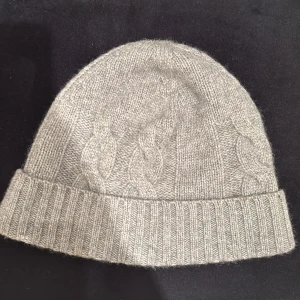 Grå kashmirmössa från John Henric - Snygg och mjuk grå mössa i kashmir från John Henric. Den har ett klassiskt flätmönster och ribbad kant som ger en stilren look. Perfekt för kyliga dagar. Nypris 799kr
