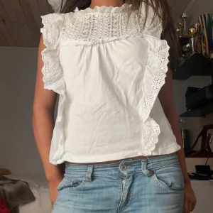 Vit blus med spetsdetaljer - Säljer en söt vit blus med spetsdetaljer och volanger på ärmarna. Blusen har en lös passform och är perfekt för en somrig look. Den är tillverkad i ett lätt och luftigt material.