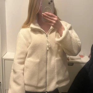 Beige teddyjacka med dragkedja - Mysig beige teddyjacka med dragkedja och hög krage. Perfekt för kyliga dagar med sin fluffiga och varma design. Jackan har långa ärmar och två praktiska fickor framtill.