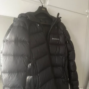 Svart dunjacka från Peak Performance - Snygg och varm svart dunjacka från Peak Performance. Jackan har en dragkedja framtill och en praktisk huva. Perfekt för kyliga dagar.