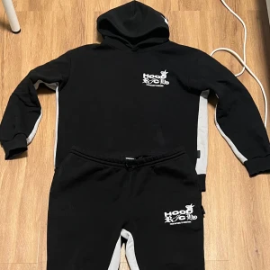 Svart och grå hoodie med byxor från Hoodrich - Säljer en svart och grå hoodie med matchande byxor från Hoodrich. Setet har en cool logga på bröstet och benet. Hoodien har en klassisk design med huva och långa ärmar, medan byxorna har en bekväm passform med elastisk midja.