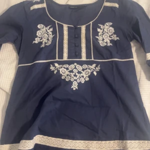 Mörkblå blus med broderade blommor  - Säljer en mörkblå blus från Kappahl med vackra vita broderade blommor och spetsdetaljer. Priset kan diskuteras 