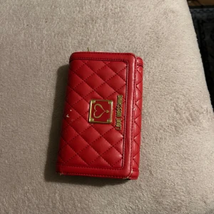 Röd quiltad plånbok från Love Moschino - Snygg röd plånbok från Love Moschino med quiltat mönster och gulddetaljer. Den har en elegant design med ett hjärta på framsidan och praktisk dragkedja.