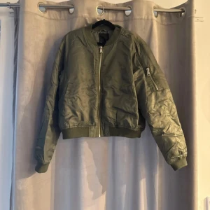 Grön bomberjacka med orange foder - Snygg grön bomberjacka med dragkedja och orange foder. Jackan har en klassisk bomberstil med ribbade muddar och en praktisk ficka med dragkedja på ärmen. Perfekt för en cool och avslappnad look. Passar mig som har storlek L jackor 