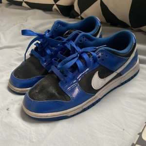 Nike Dunk i blått och svart - Snygga Nike Dunk sneakers i blått och svart med vit swoosh. Skorna har en klassisk design med snörning och en slitstark sula. Perfekta för en sportig look.