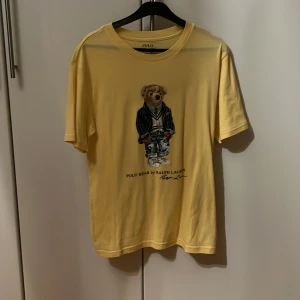 Gul t-shirt med Polo Bear från Ralph Lauren - Säljer en gul t-shirt från Ralph Lauren med det ikoniska Polo Bear-motivet på framsidan. T-shirten har en normal passform och är kortärmad, perfekt för en avslappnad stil. Ändvänd fåtal gånger. Och är i banstorlek 160/80 som passar 14 till 16 åringar.