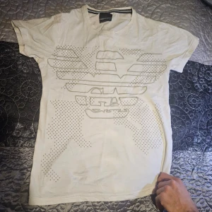 Vit t-shirt från Emporio Armani - Snygg vit t-shirt från Emporio Armani med ett stilrent mönster på framsidan. T-shirten är kortärmad och har en normal passform. Perfekt för en avslappnad och trendig look. Man S, L kvinna skulle jag vilja säga.