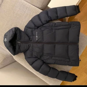 Svart dunjacka med huva - Snygg svart dunjacka från sail racing med huva och dragkedja. Perfekt för kalla dagar med sin pufferdesign som håller dig varm. Jackan har långa ärmar och en stilren look.