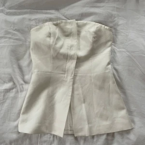 Vit axelbandslös topp - Snygg vit axelbandslös topp med en stilren design. Perfekt för en elegant look. Toppen har en rak skärning och en diskret slits framtill.