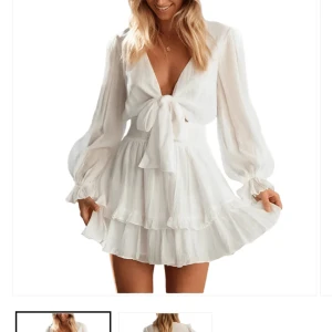 Vit klänning från Dressy - Vit kortklänning med volangdetaljer och knyt framtill. Klänningen har långa ärmar och en djup v-ringning, perfekt till studenten. Prislapp kvar! 