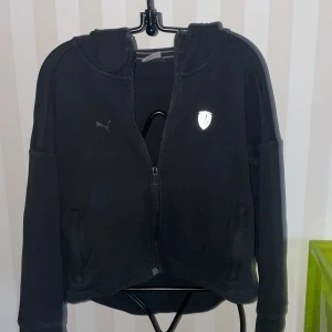 Svart huvtröja från Puma - Säljer en svart huvtröja från Puma med dragkedja och Ferrari-logga på bröstet. Tröjan har långa ärmar och en bekväm passform, perfekt för vardagsbruk. Huvan ger extra stil och funktionalitet.