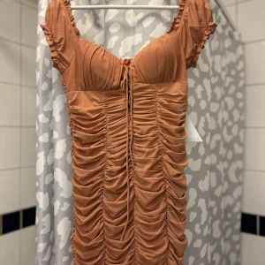 Brun klänning från Love Triangle - Snygg brun klänning från Love Triangle med rynkade detaljer och korta ärmar. Den har en åtsittande passform och en söt knytning framtill. Perfekt för en stilfull look.