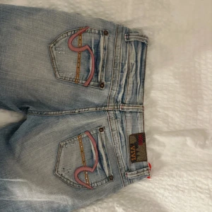 Ljusblå jeans med broderade detaljer - Säljer ett par ljusblå jeans från miss Vivi, med unika broderade detaljer på bakfickorna i rosa och orange. Jeansen har en bootcut-stil och är lågmidjade med en klassisk femficksdesign. Perfekta för en avslappnad look! Lågmidjade bootcut utsvängda low rise unika. Skulle säga att de passar någon lite kortare.