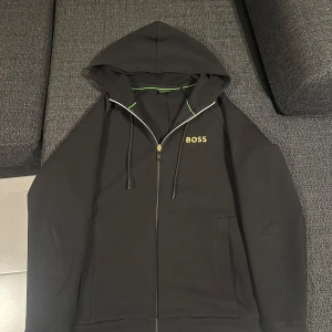 Svart hoodie från Boss - Snygg svart hoodie från Boss med dragkedja och huva. Den har en stilren design med logotypen i gult på bröstet. Perfekt för en avslappnad stil. Aldrig använt pågrund av fel storlek. 