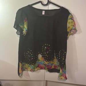 Svart blus med färgglada detaljer - Säljer en svart blus med korta ärmar och färgglada blommiga detaljer. Blusen har ett unikt mönster med utskärningar och en lös passform. Perfekt för att liva upp garderoben!