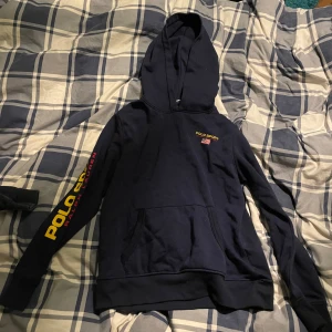 Mörkblå hoodie från Polo Ralph lauren Sport - Säljer en mörkblå hoodie från Polo Ralph lauren Sport med tryck på ärmen och bröstet. Tröjan har en klassisk känguruficka och en bekväm huva. Perfekt för en avslappnad stil.