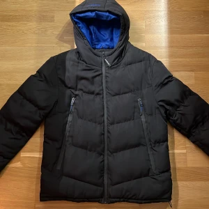 Svart dunjacka från Closure LDN - Snygg svart dunjacka från Closure LDN med blått foder. Jackan har en huva och dragkedja framtill samt två praktiska fickor med dragkedja. Perfekt för kyliga dagar. Gratis, ni står för frakten. 
