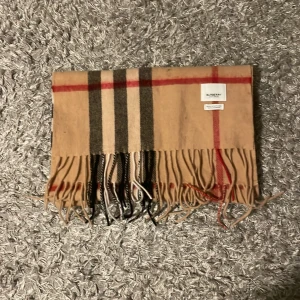 Beige rutig halsduk från Burberry - Säljer en klassisk beige rutig halsduk från Burberry, tillverkad i Skottland av 100% kashmir. Den har ett tidlöst rutmönster i svart, rött och vitt med fransar i ändarna. Perfekt för att hålla dig varm och stilren under kyliga dagar.
