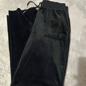 Svarta mjukisbyxor från Juicy Couture - Snygga svarta mjukisbyxor från Juicy Couture i mjukt material. Byxorna har en elastisk midja med snörning och praktiska fickor både fram och bak. Perfekta för en avslappnad stil. ( ner kortade i benen) 