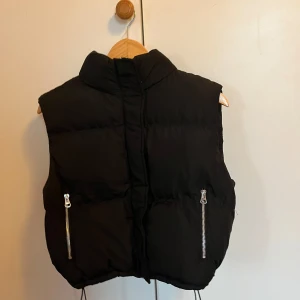 Svart dunväst med dragkedja - Snygg svart dunväst med dragkedja framtill och två praktiska fickor med dragkedjor. Perfekt för kyligare dagar och enkel att matcha med olika outfits.