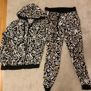 Svart och vit hoodie med mönster - Cool tracksuit från Moschino i svart och vitt med ett unikt mönster av bokstäver och små figurer. Den har en dragkedja framtill och en bekväm huva med snörning. Perfekt för en avslappnad stil eller en chill dag hemma. Möts i sthlm!