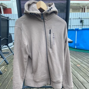 Beige hoodie från Sail Racing - Säljer en snygg beige hoodie från Sail Racing i bra skick. Den har en dragkedja framtill och en praktisk bröstficka med dragkedja. Perfekt för höst och vår, med en skön huva och långa ärmar. Passar både till vardags och lite mer avslappnade tillfällen. Knappt använd och priset ej hugget i sten