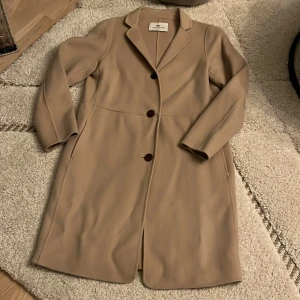 Beige kappa från DAY - Säljer en elegant beige kappa från DAY i mycket bra skick. Den har en klassisk design med knappar framtill och långa ärmar. Ger en stilren look till vilken outfit som helst. Passar både till vardags och festliga tillfällen.