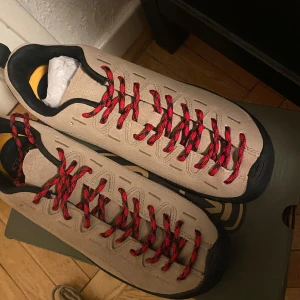 Beige sneakers med röda snören - Säljer ett par snygga beige sneakers med röda och svarta snören. Skorna har en robust design med svart sula och detaljer. Perfekta för både vardag och äventyr. De är i mycket bra skick och redo för nya äventyr!