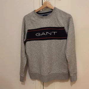 Gant tröja - Säljer en snygg grå sweatshirt från GANT i mycket bra skick. Den har ett stilrent tryck med GANT-loggan i vitt och rött på bröstet. Perfekt för både vardag och chill. Nypris: 1200