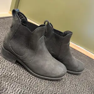 Säljer ett par snygga svarta boots från UGG i mycket bra skick. De är gjorda i skinn och har en klassisk chelsea boots-stil med elastiska paneler på sidorna för enkel på- och avtagning. Perfekta för höst och vinter!