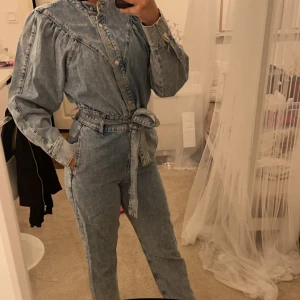 Jeansjacka med knytband - Säljer en cool jeansjacka i ljusblå denim med knappar och knytband i midjan. Jackan har långa ärmar och en unik design med puffiga axlar. Perfekt för en avslappnad men stilren look. Passar bra till både vardag och fest!