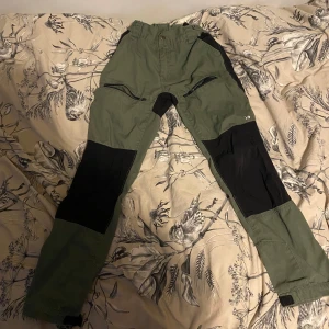 Gröna och svarta friluftsbyxor - Säljer ett par gröna och svarta friluftsbyxor i mycket bra skick. De har en praktisk design med dragkedjor och förstärkta knän, perfekt för utomhusaktiviteter. Byxorna har en normal passform och är gjorda av slitstarkt material. Perfekta för vandring eller andra äventyr i naturen! 