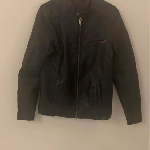 Svart skinnjacka med dragkedja - Säljer en snygg svart skinnjacka i bra skick. Den har en dragkedja framtill och en cool bröstficka med dragkedja. Perfekt för höst och vår, och ger en stilren look. Passar både till vardags och fest!