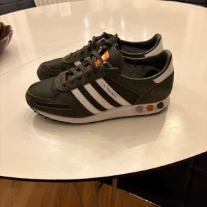Adidas LA Trainer i olivgrön - Säljer ett par snygga Adidas LA Trainer sneakers i olivgrön färg. Skorna har använts men är fortfarande i ett väldigt bra skick. Skorna har liten defekt inuti men är verkligen inget man märker av. Storlek 44 2/3 och box tillkommer