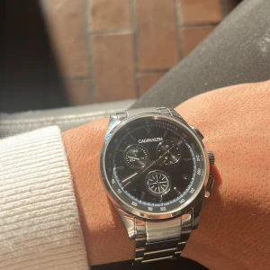 Calvin Klein herrklocka i silver - Snygg Calvin Klein herrklocka i silver med svart urtavla och tre mindre urtavlor för extra funktioner. Armbandet är i rostfritt stål och klockan har en stilren design som passar både till vardags och fest. Perfekt accessoar för att lyfta din stil!