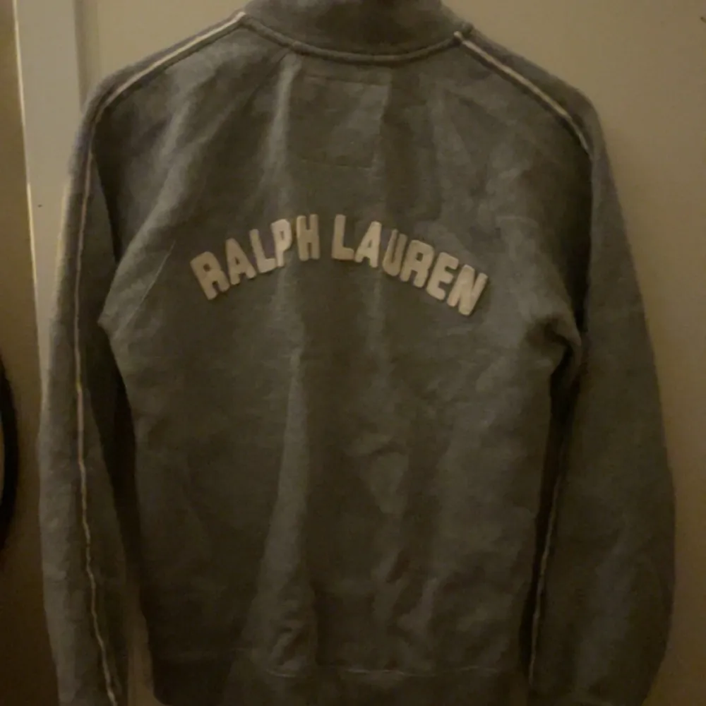 Säljer en snygg grå tröja från Ralph Lauren med dragkedja och hög krage. Den har texten 'POLO JEANS COMPANY' på framsidan och 'RALPH LAUREN' på baksidan. Perfekt för en casual look eller en chill dag hemma. Passar bra till jeans eller joggers!. Tröjor & Koftor.