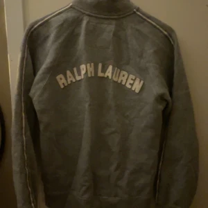 Grå tröja från Ralph Lauren - Säljer en snygg grå tröja från Ralph Lauren med dragkedja och hög krage. Den har texten 'POLO JEANS COMPANY' på framsidan och 'RALPH LAUREN' på baksidan. Perfekt för en casual look eller en chill dag hemma. Passar bra till jeans eller joggers!
