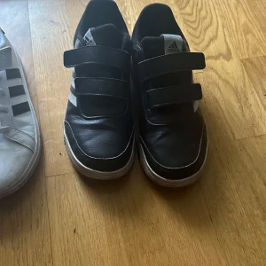 Svarta Adidas sneakers med kardborreband - Säljer ett par svarta Adidas sneakers i bra skick. De har tre praktiska kardborreband för enkel på- och avtagning. Perfekta för vardagsbruk och passar till det mesta. Skorna har en vit sula och de klassiska tre ränderna i vitt på sidan.