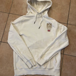 Vit hoodie från Morris - Säljer en stilren vit hoodie från Morris med broderad logga och brittisk flagga på bröstet. Den har en klassisk känguruficka och justerbar huva med snören. Perfekt för en avslappnad stil eller kyliga dagar. Passar både till vardags och lite finare tillfällen.