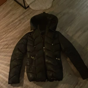Svart dunjacka från Garoff - Säljer en snygg svart dunjacka från Garoff i storlek 34. Jackan har en mysig huva och ett coolt leopardmönstrat foder. Den är perfekt för kalla dagar med sin quiltade design och dragkedja. Dessutom medföljer ett bälte för en mer figurnära passform. Perfekt för höst och vinter!