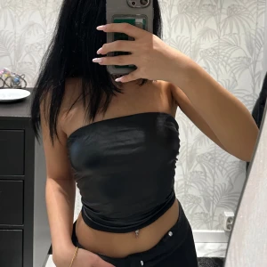 Svart tubtopp - Snygg svart tubtopp i glansigt satinmaterial. Perfekt för en utekväll eller fest! Toppen är axelbandslös och har en tight passform som framhäver figuren. Den är helt oanvänd!