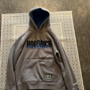 Grå hoodie från Hoodrich - Säljer en snygg grå hoodie från Hoodrich i storlek S. Den har en stor ficka fram och texten 'Hoodrich' i svart och blått på bröstet. Insidan av huvan är blå och den har en mjuk insida för extra komfort. Perfekt för kyliga dagar!