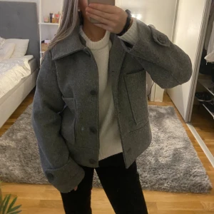 Grå kort kappa  - Säljer en snygg grå kappa från Kappahl. Den har en klassisk design med stora knappar och två fickor framtill. Perfekt för höst och vår. Tyget har blivit nopprigt (se bild 4) men utöver det är den i mycket bra skick! Storlek xs 💓💓💓